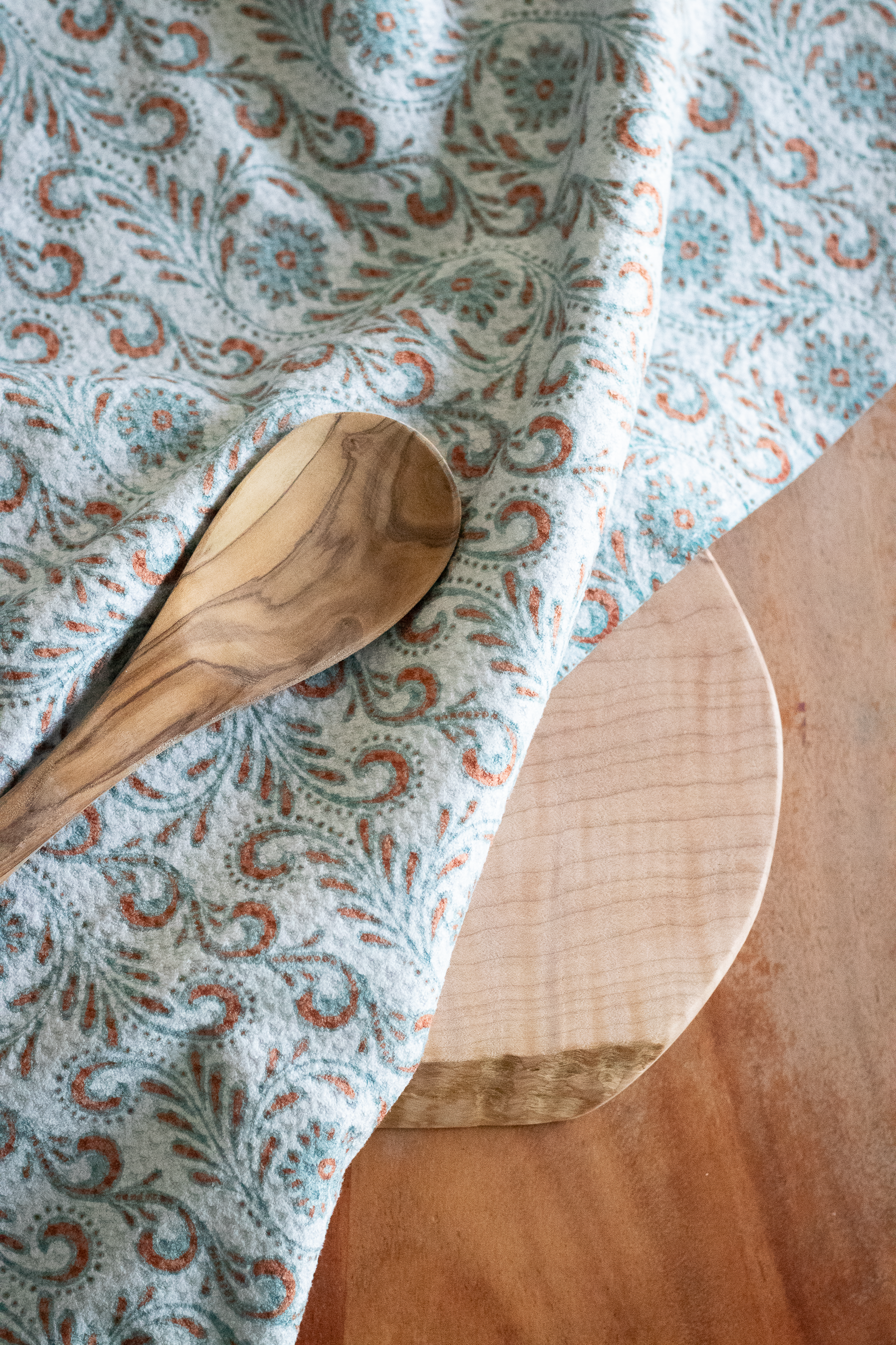 Close-up of a wooden spoon on a waffle-weave kitchen tea towel with a blue and terracotta-colored tooled leather floral design on a wooden surface.