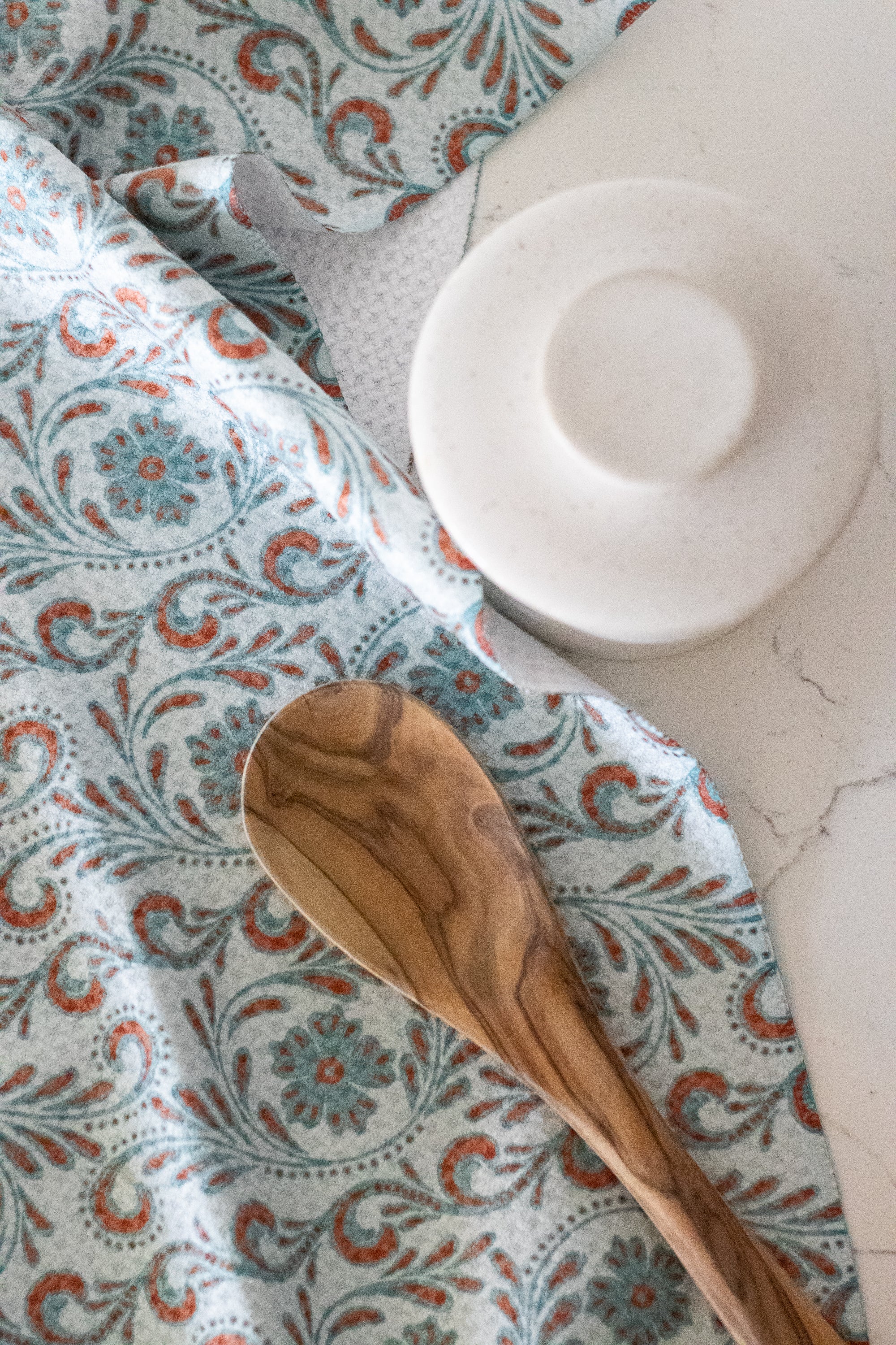 Close-up of a wooden spoon on a waffle-weave kitchen tea towel with a blue and terracotta-colored tooled leather floral design, on a white surface with a white butter bell jar.