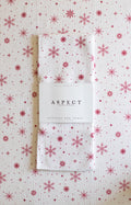 A folded waffle-weave kitchen tea towel with a red snowflake pattern on a white background, wrapped in a white paper belly band labeled 'ASPECT HOME GOODS' & 'Kitchen Tea Towel'. The towel is laid on top of another towel with the same pattern.