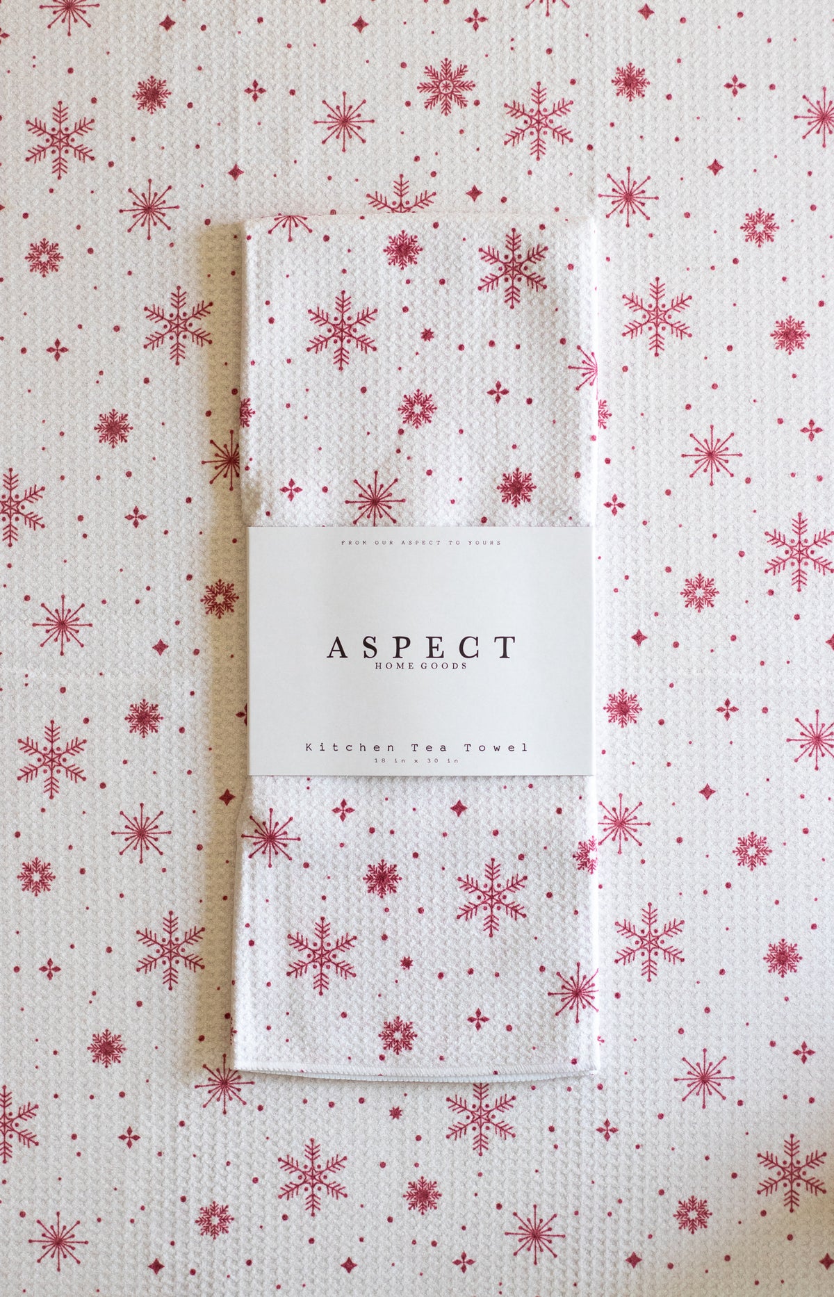 A folded waffle-weave kitchen tea towel with a red snowflake pattern on a white background, wrapped in a white paper belly band labeled 'ASPECT HOME GOODS' & 'Kitchen Tea Towel'. The towel is laid on top of another towel with the same pattern.