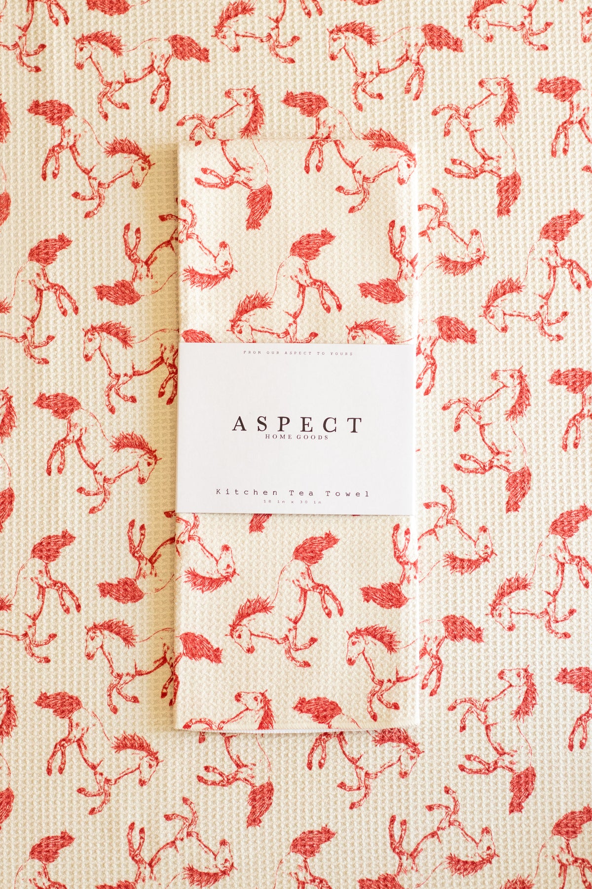 A folded waffle-weave kitchen tea towel with a red running horses pattern on a cream background, featuring a white paper belly band label in the center that reads 'ASPECT HOME GOODS' and 'KITCHEN TEA TOWEL', laid on a background of the same pattern.