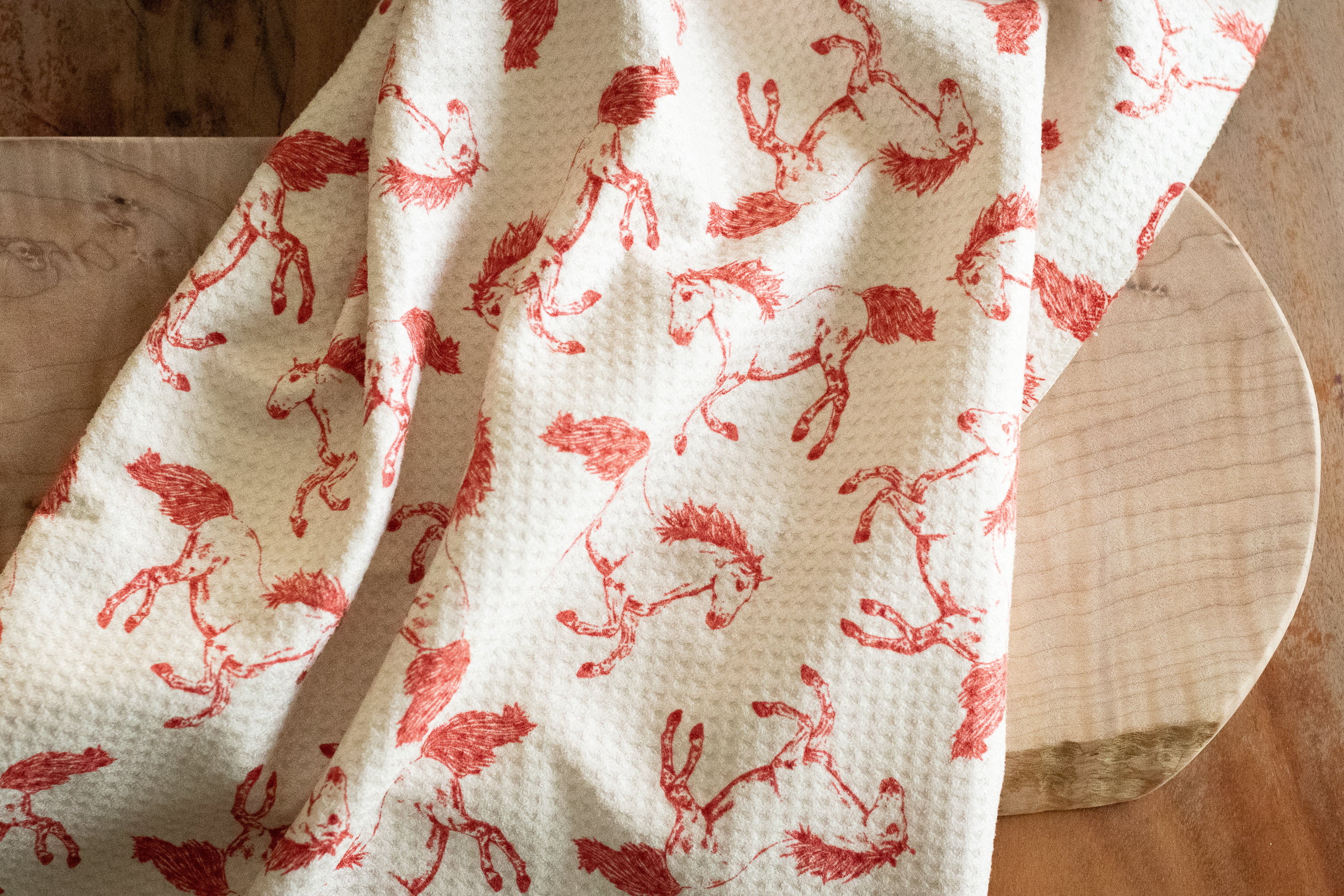 A waffle-weave kitchen tea towel with a pattern of hand-drawn red horses on a light beige background, on a wooden surface.