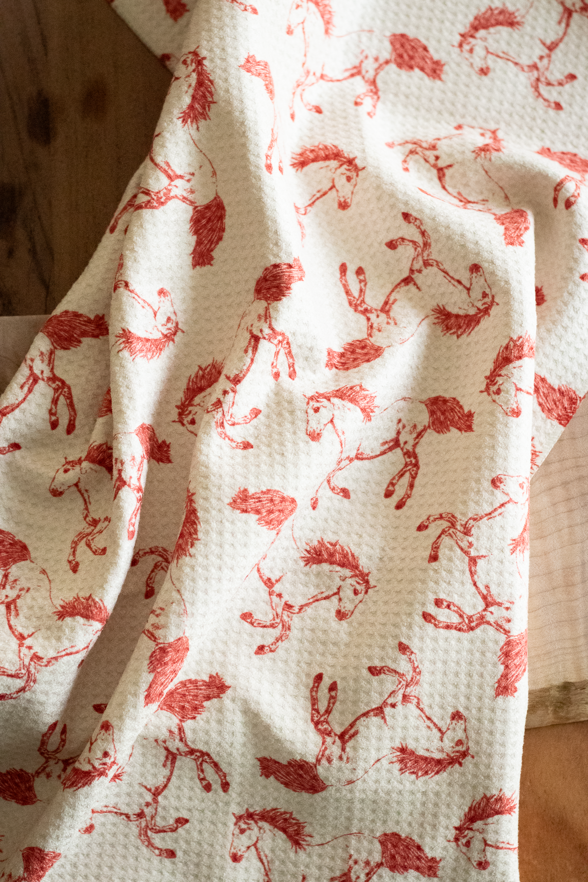 A waffle-weave kitchen tea towel with a pattern of hand-drawn red horses on a light beige background, on a wooden background.