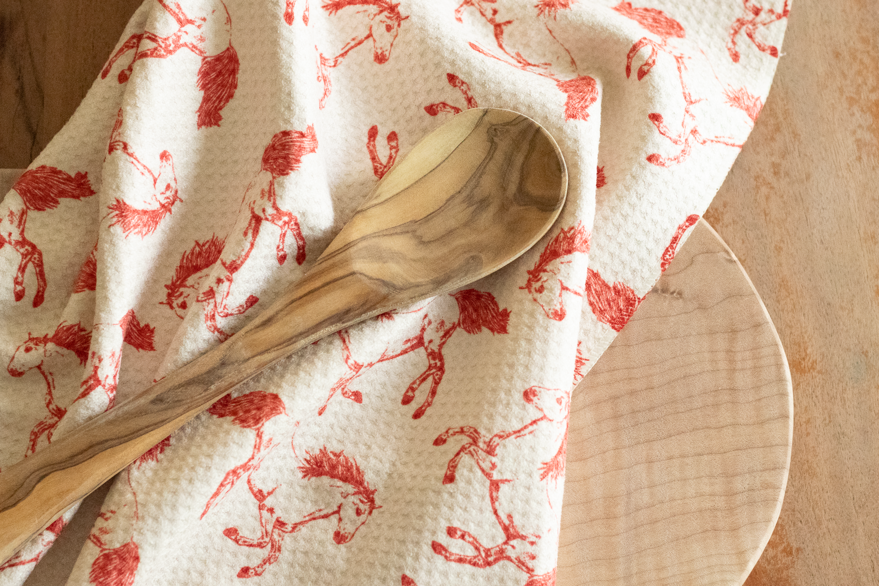 Wooden spoon on a light beige waffle-weave kitchen tea towel with a hand-drawn red horse pattern, on a wooden surface.