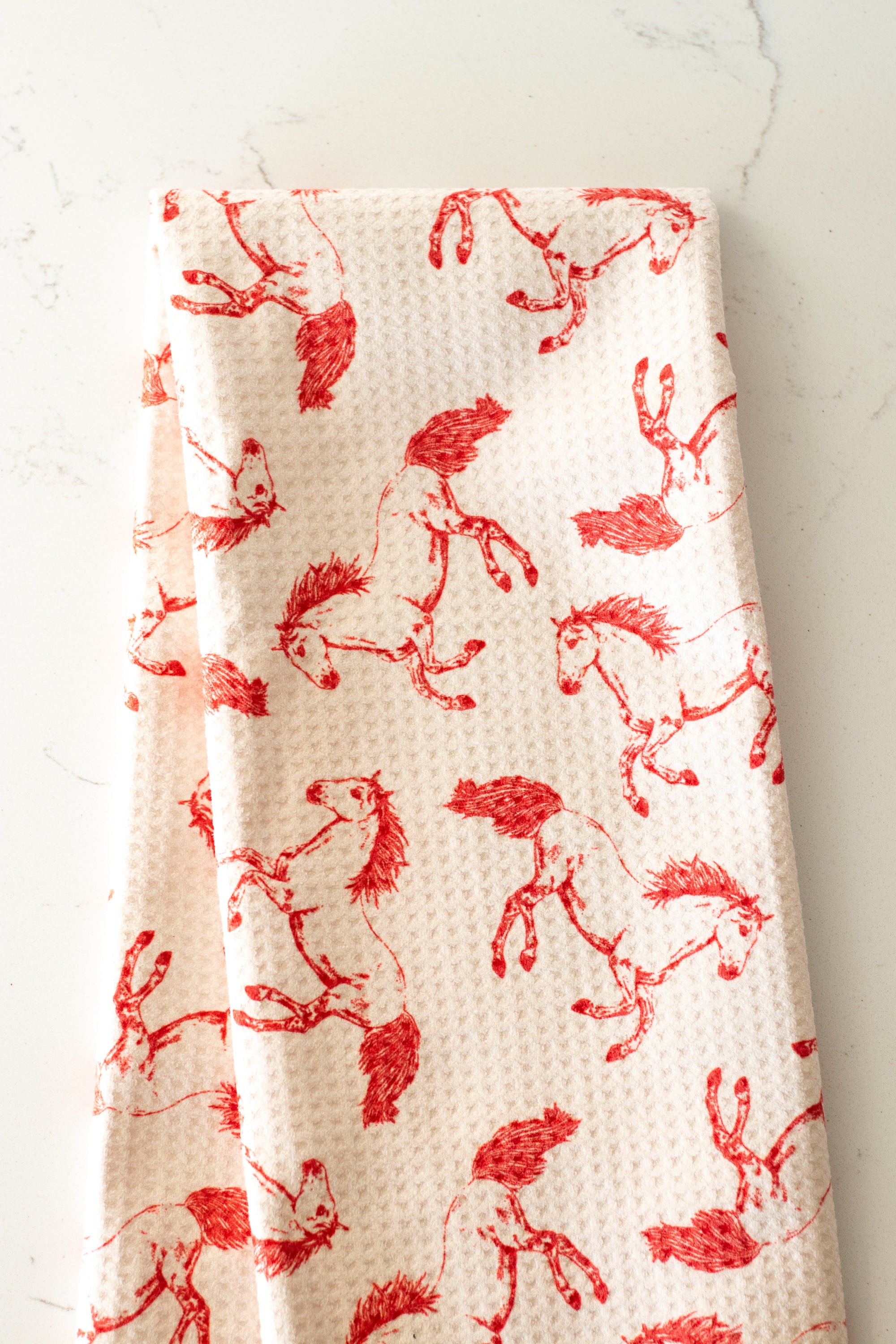 Light beige warfle-weave kitchen tea towel with a red horse pattern, folded lengthwise in half, on a white stone surface.