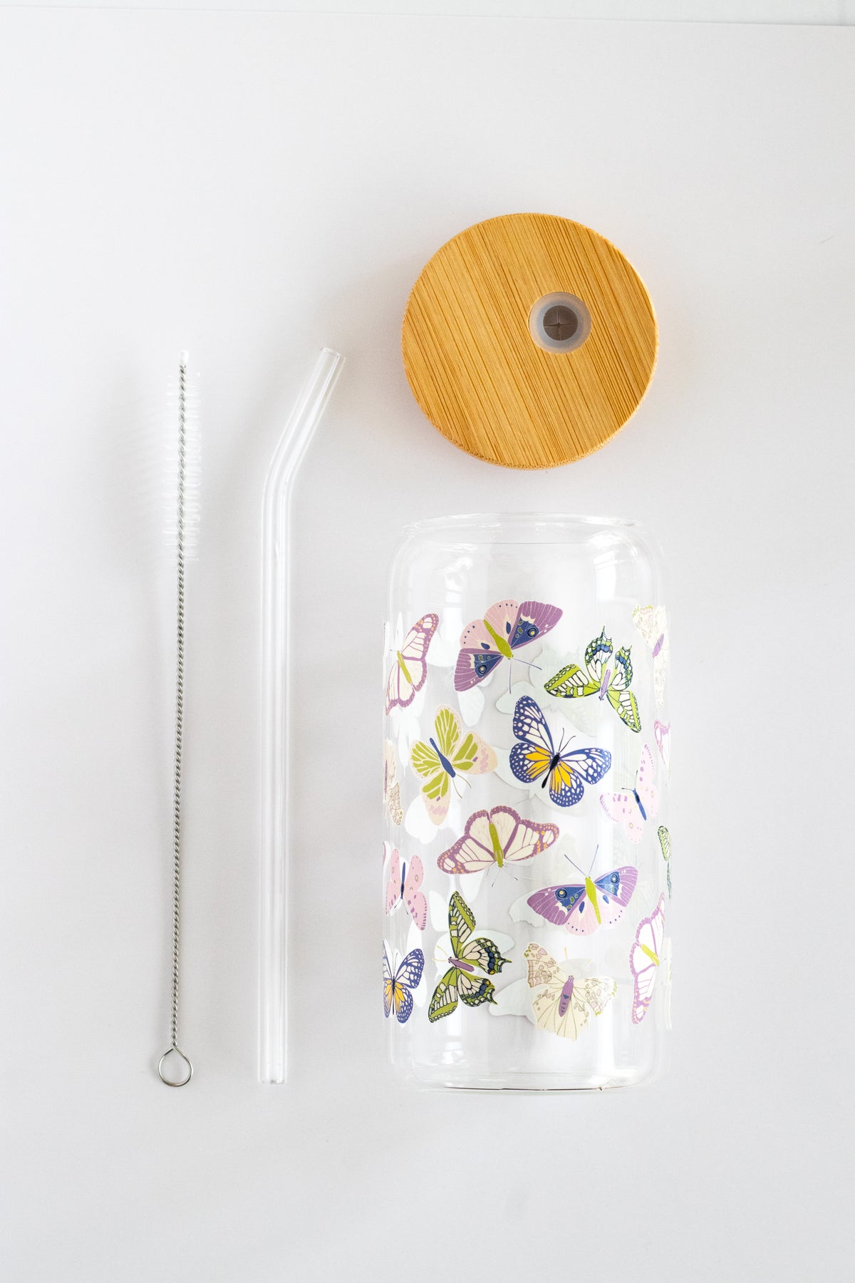 Can glass with a butterfly design, bamboo wooden lid, glass straw, and straw cleaning brush, on a white background.
