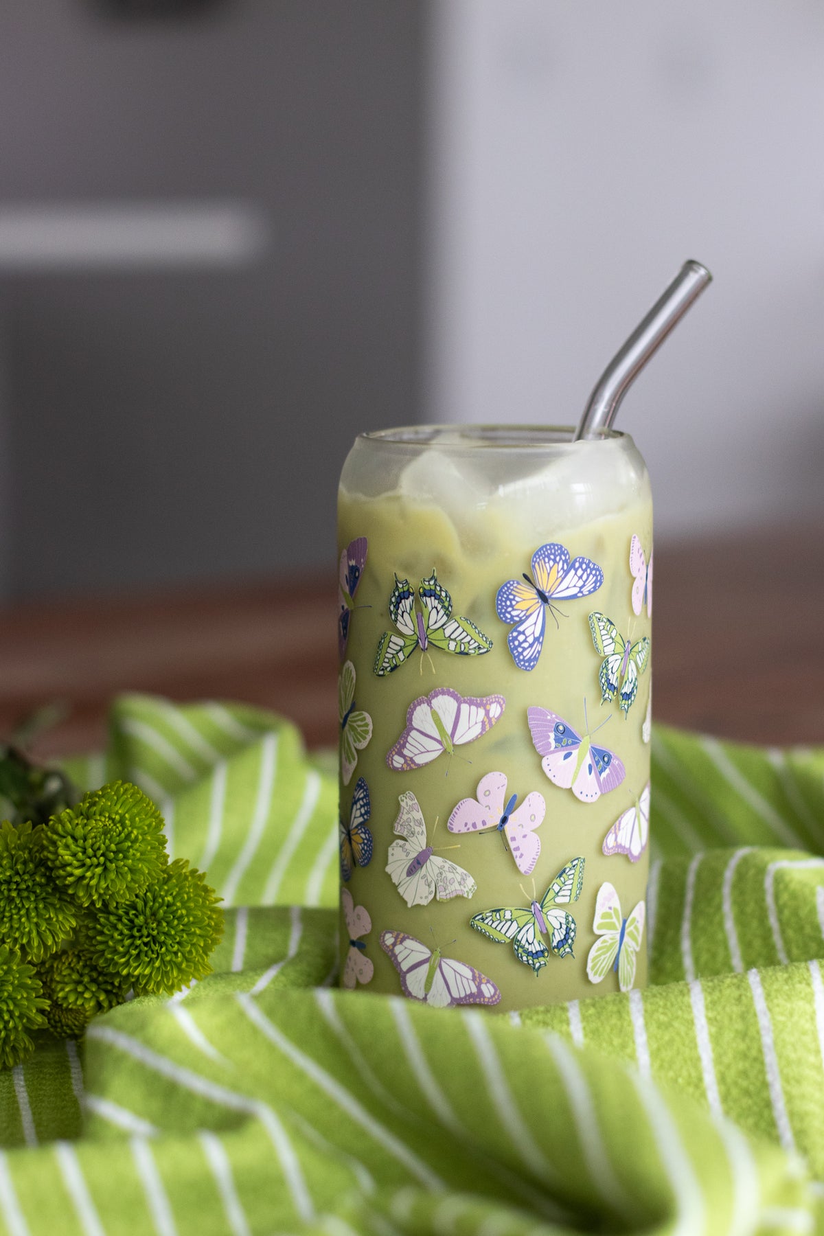 Can glass with a butterfly design filled with a matcha latte on a striped bright green and white waffle-weave kitchen tea towel with green flowers.