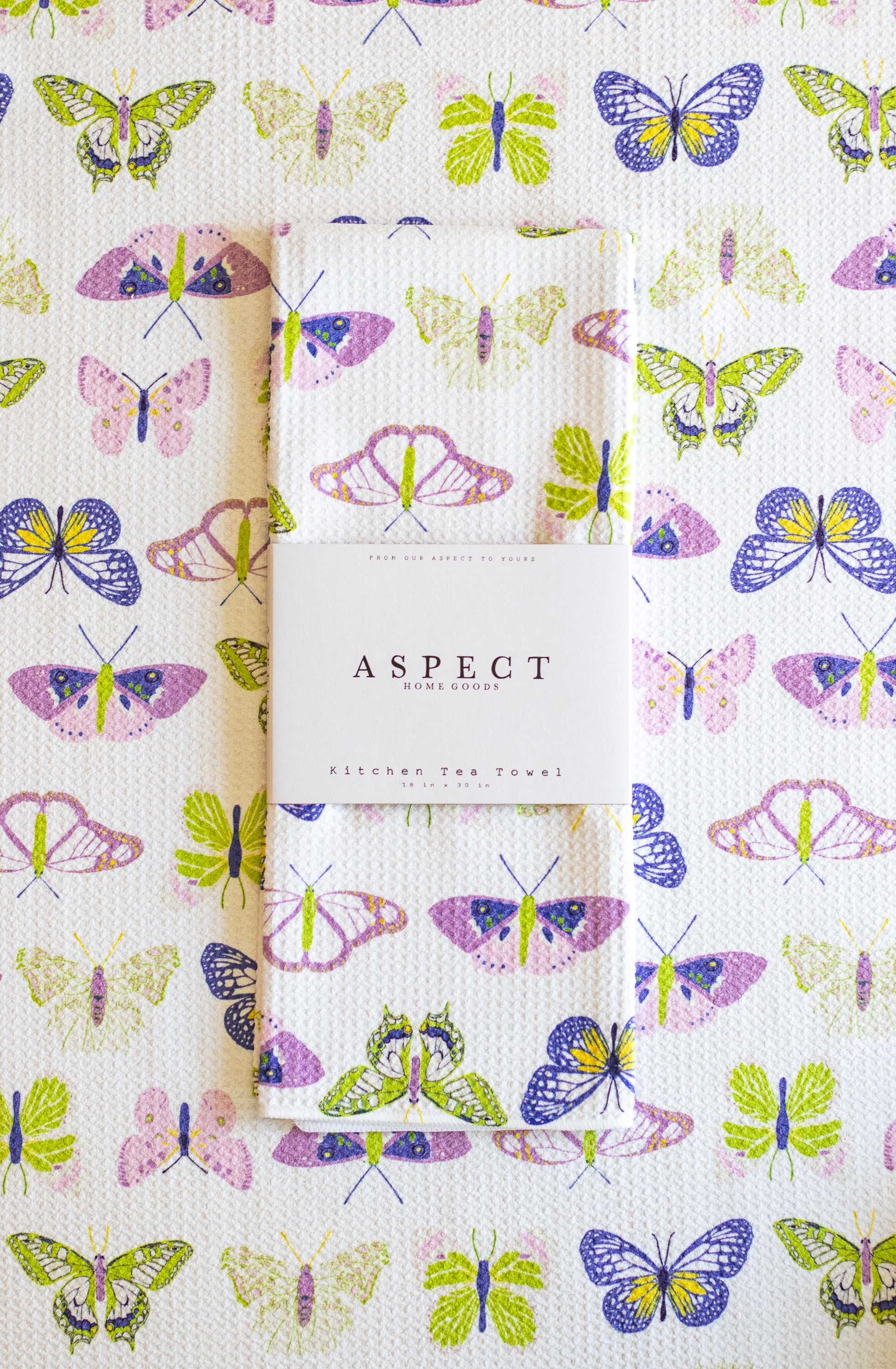 A folded white kitchen Tea Towel with a colorful butterfly print in lime green, indigo and purple, wrapped in a white paper belly band labeled 'ASPECT HOME GOODS',laid flat on a matching butterfly-patterned background.