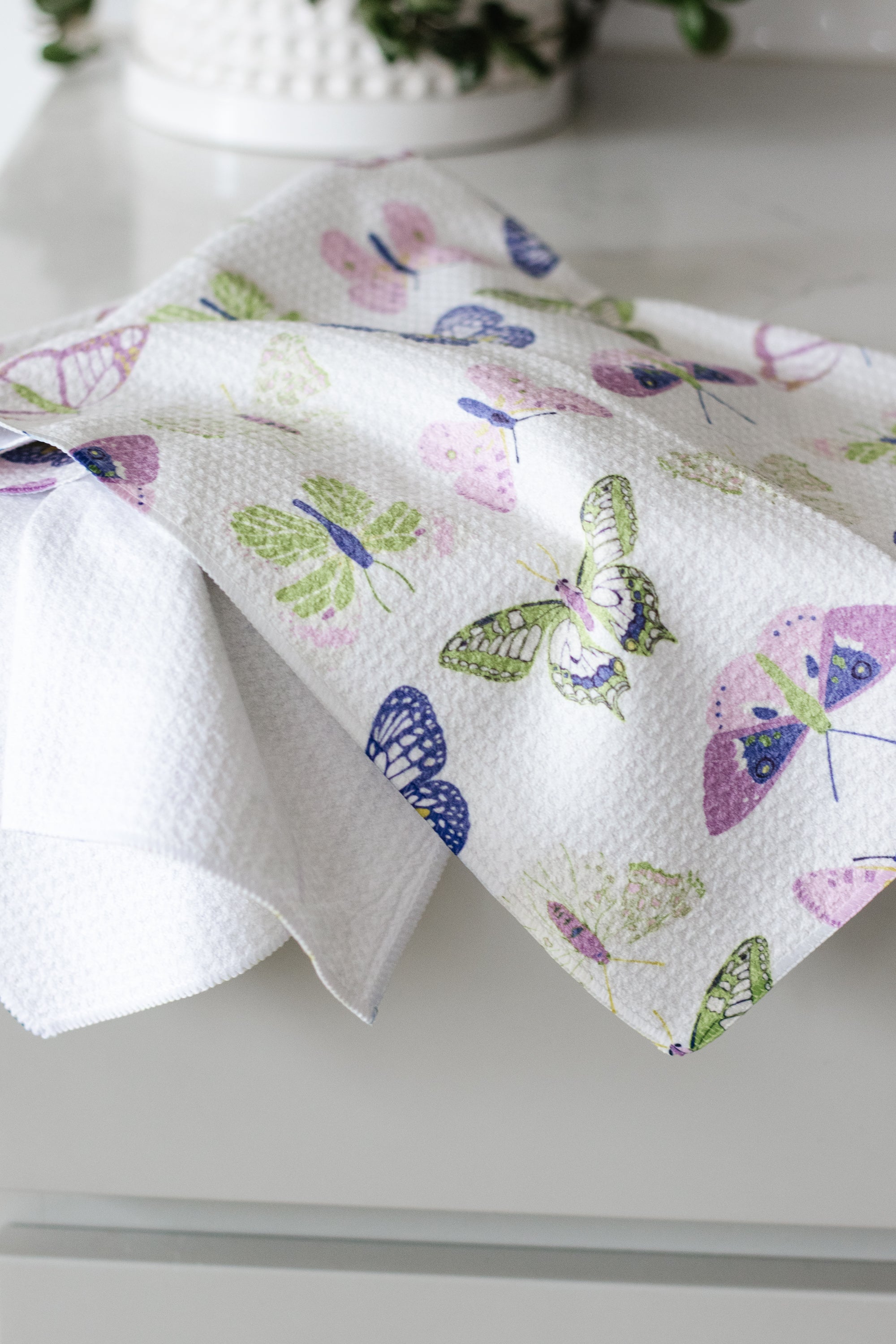 Butterfly-patterned waffle-weave kitchen tea towel on a white countertop with a plant in the background. 