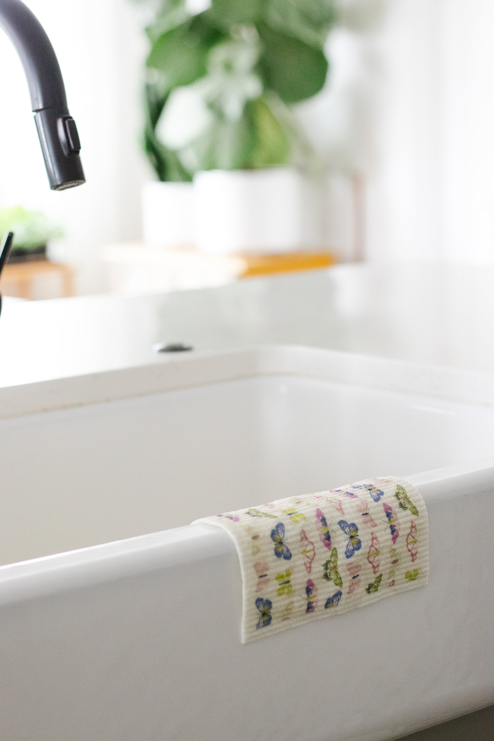 A Swedish dishcloth with a butterfly pattern draped over the edge of a white farmhouse kitchen sink.