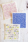 Three Swedish dishcloths are arranged on a distressed white wooden surface. One napkin has a pink and purple floral pattern, one has a blue background with small white floral motifs, and one has a white background with colorful butterflies and floral prints.