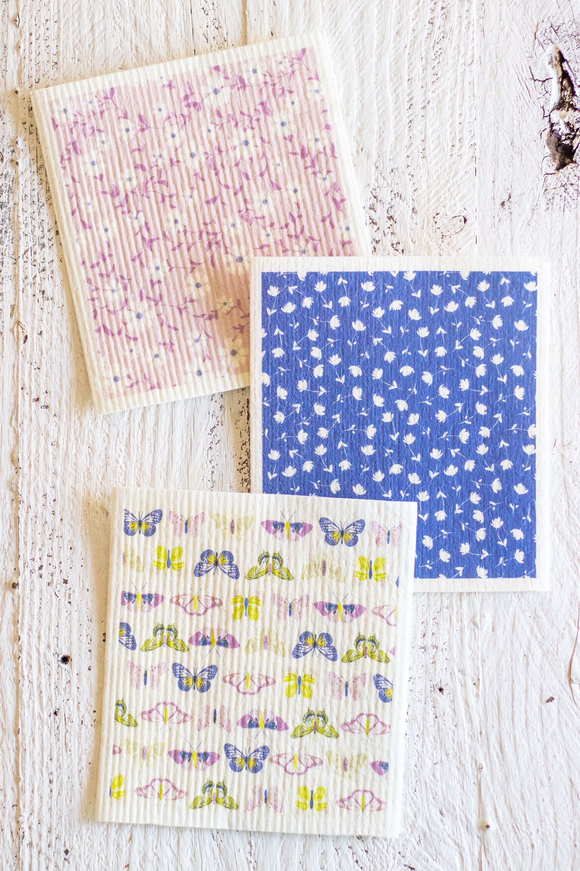 Three Swedish dishcloths are arranged on a distressed white wooden surface. One napkin has a pink and purple floral pattern, one has a blue background with small white floral motifs, and one has a white background with colorful butterflies and floral prints.
