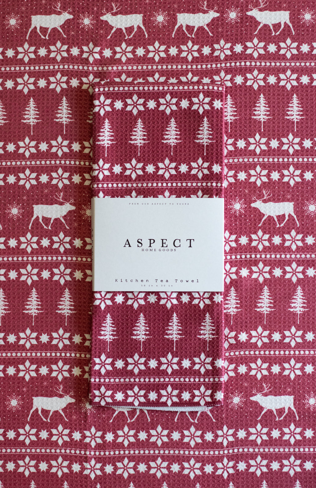A folded waffle-weave kitchen tea towel with a festive Christmas pattern, featuring white reindeer, snowflakes, and pine trees on a red background. A white paper belly band label from the brand ASPECT HOME GOODS is wrapped around the center. The towel is laid on another towel with the same pattern.