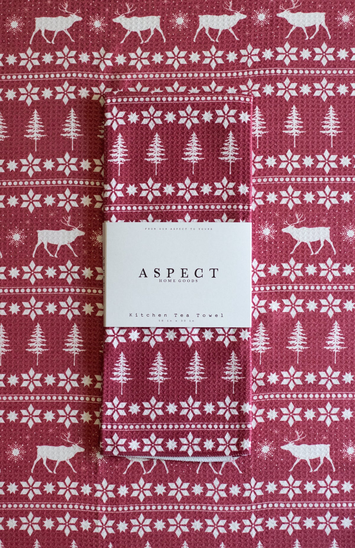 A folded waffle-weave kitchen tea towel with a festive Christmas pattern, featuring white reindeer, snowflakes, and pine trees on a red background. A white paper belly band label from the brand ASPECT HOME GOODS is wrapped around the center. The towel is laid on another towel with the same pattern.